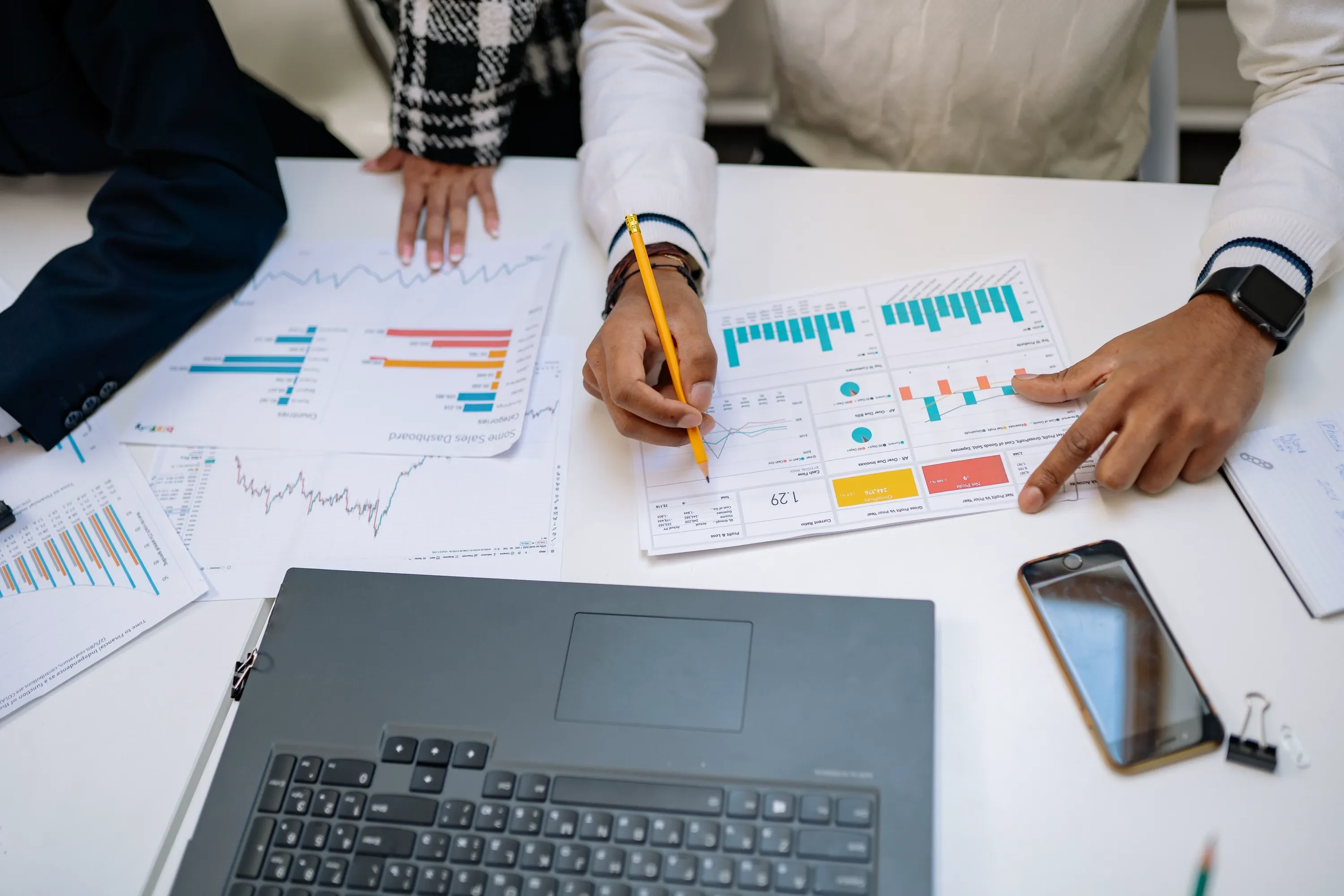 Colleagues' hands over a table of printed charts and papers, one marking a chart with a pencil beside an open laptop